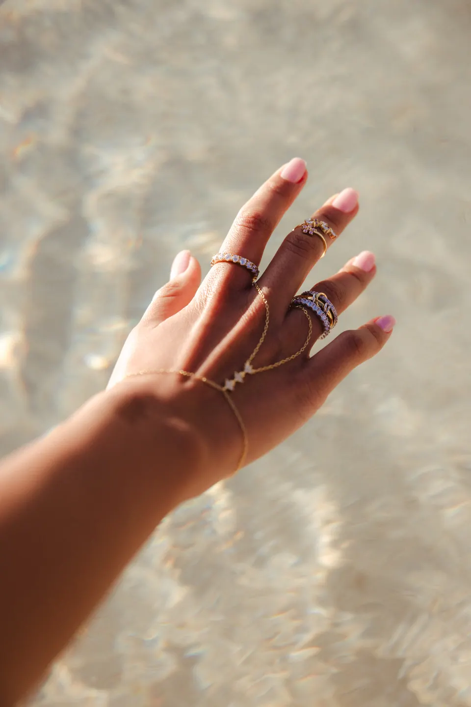 A hand reaching over rippling, sunlit water, wearing stacked rings and a hand chain.