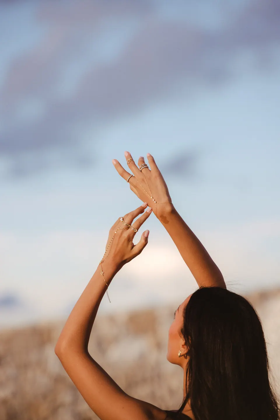 A woman seen from behind with her arms raised against a pale sky, wearing rings and a hand chain.