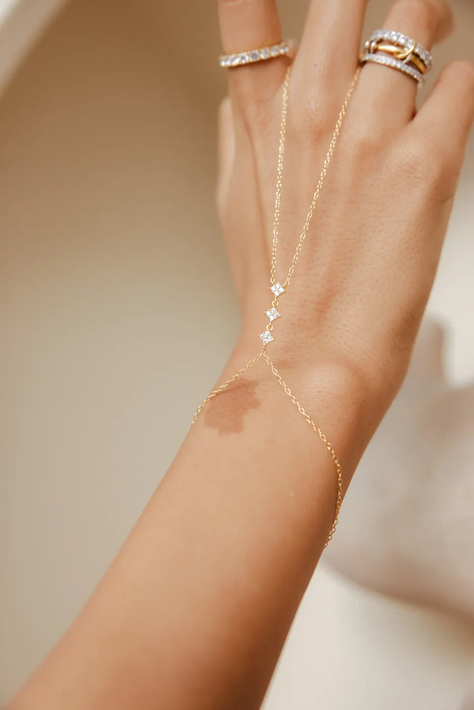 A raised hand against a cream wall, a fine hand chain running from the rings down to the wrist.
