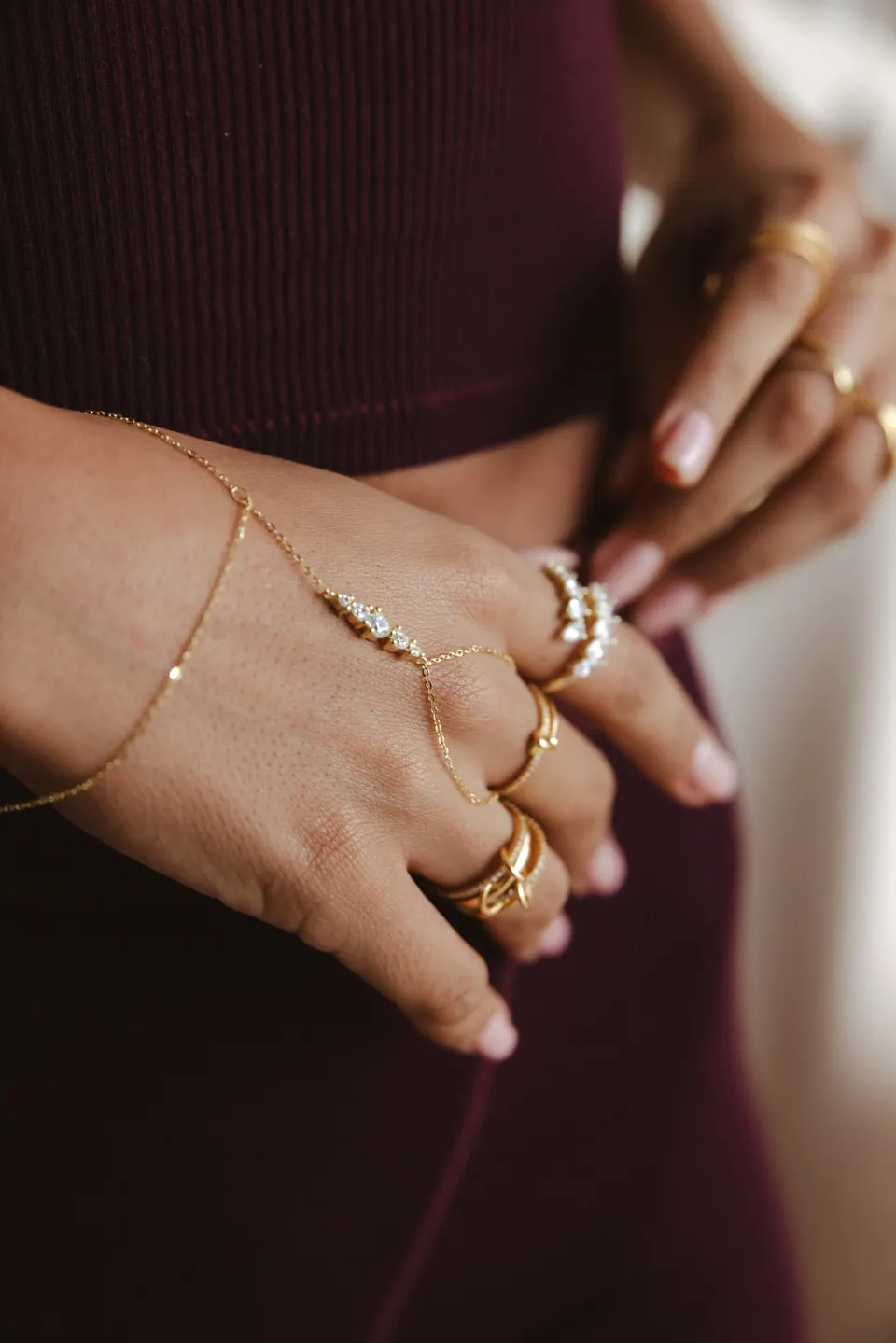 Hands at the waist of a dark burgundy outfit, wearing stacked rings and a fine hand chain.
