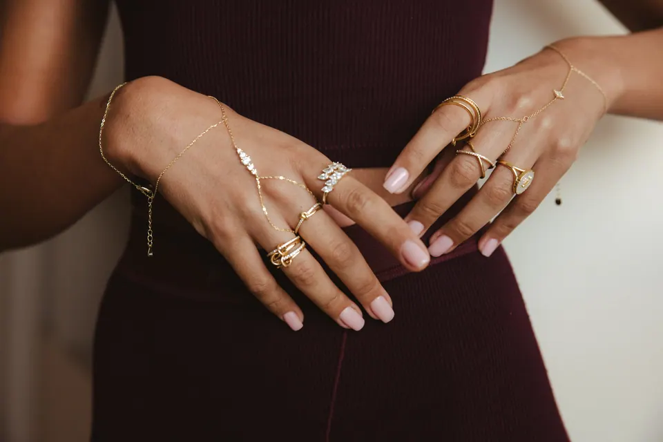 Two hands resting on the waist of a dark burgundy outfit, wearing several rings and hand chains.