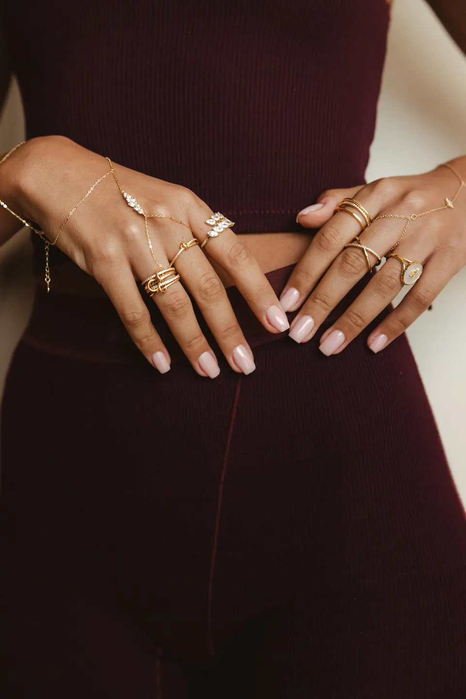 Hands resting on the waist of a burgundy knit outfit, wearing rings and hand chains, in soft window light.