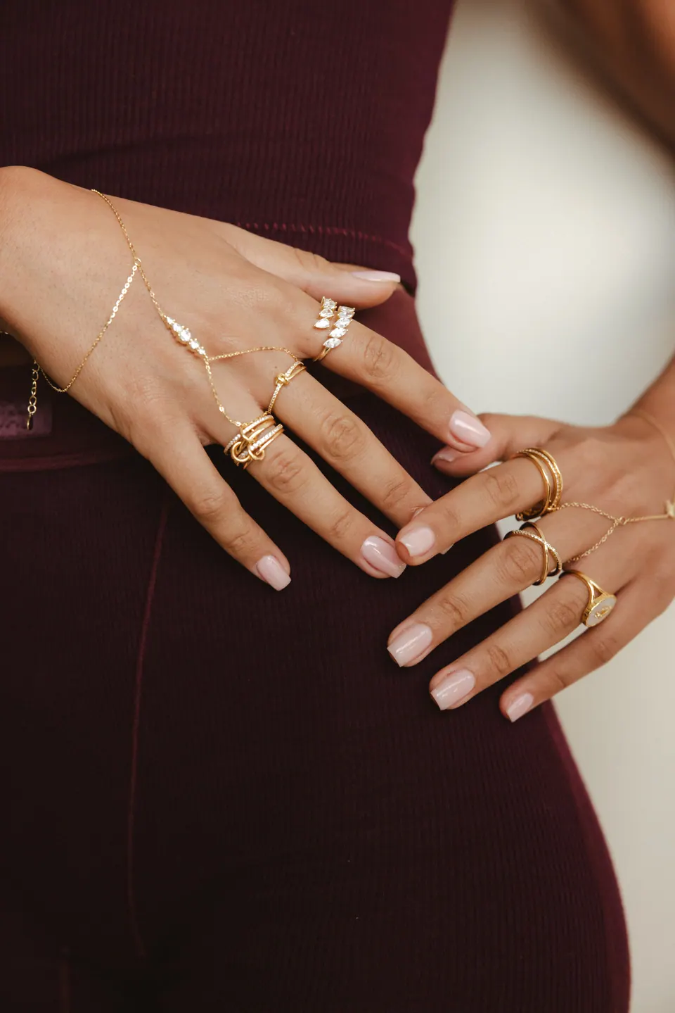 Two hands on a burgundy knit waistband, wearing several gold-plated rings and a hand chain.