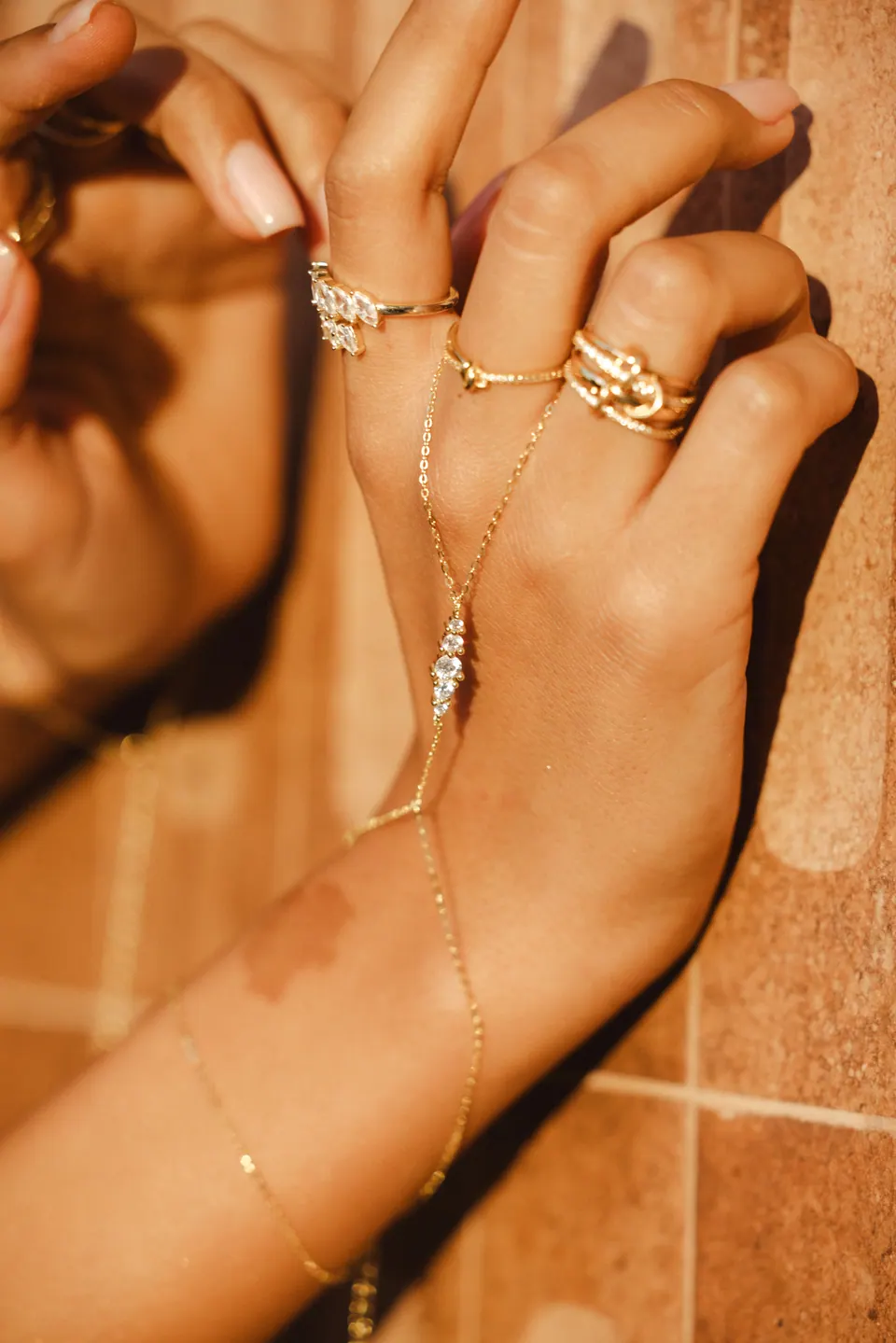 A hand with a fine hand chain raised against warm terracotta tiles in low sun.