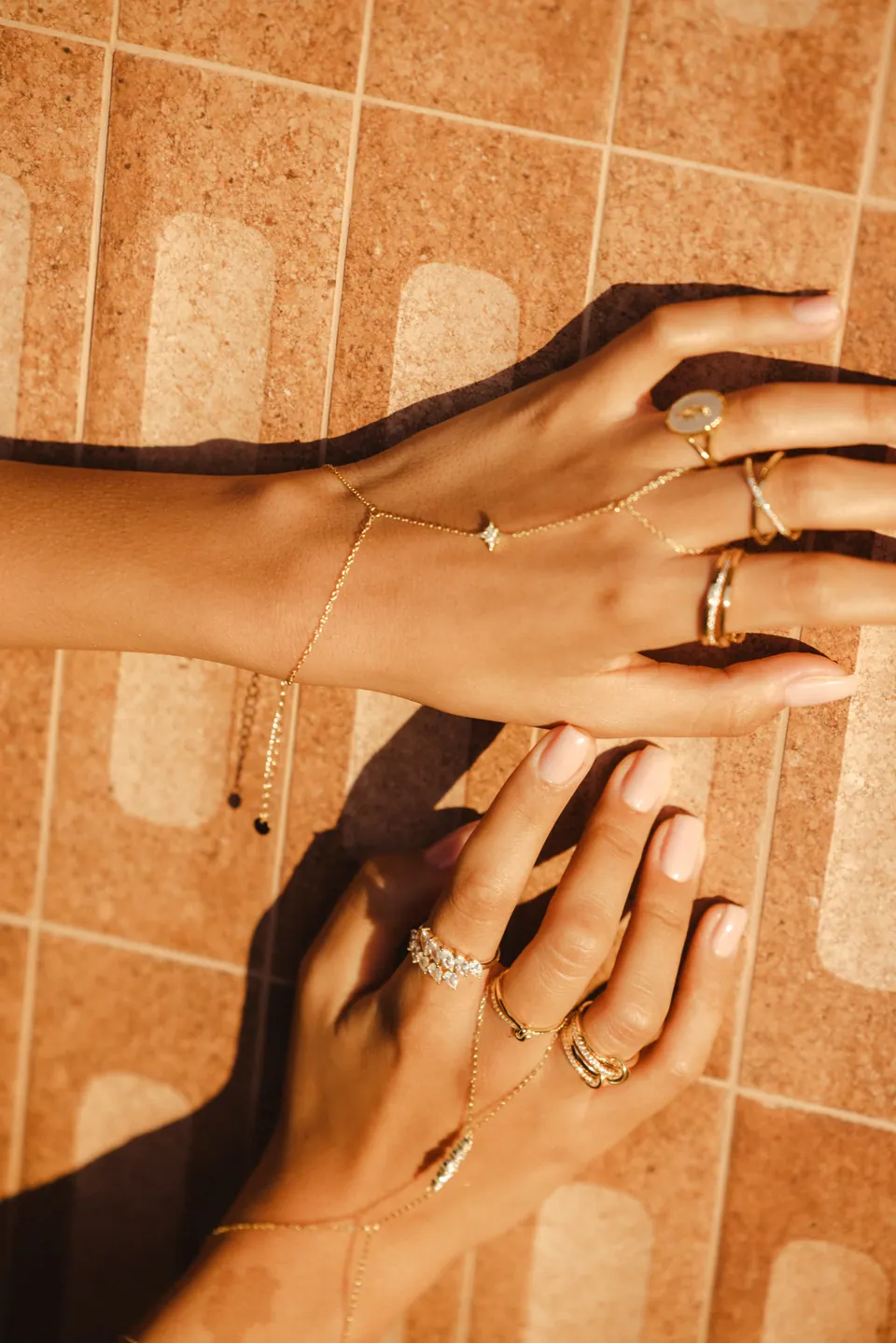 Two hands on terracotta tiles in strong sun, wearing rings and hand chains, casting sharp shadows.