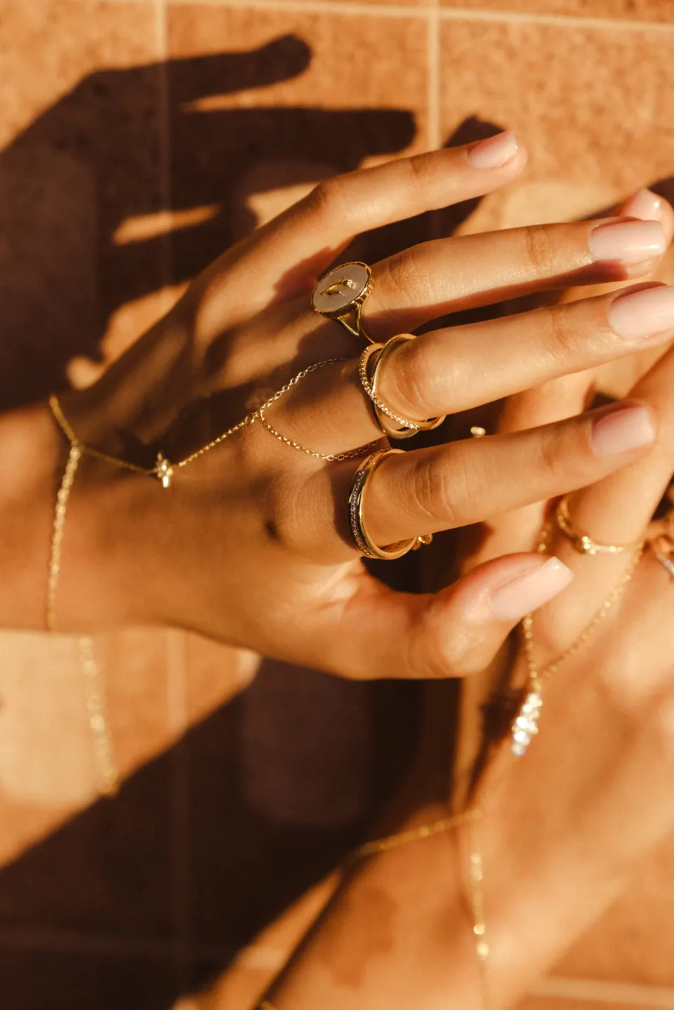 Hands and their shadows against sunlit terracotta tiles, wearing rings and a hand chain.