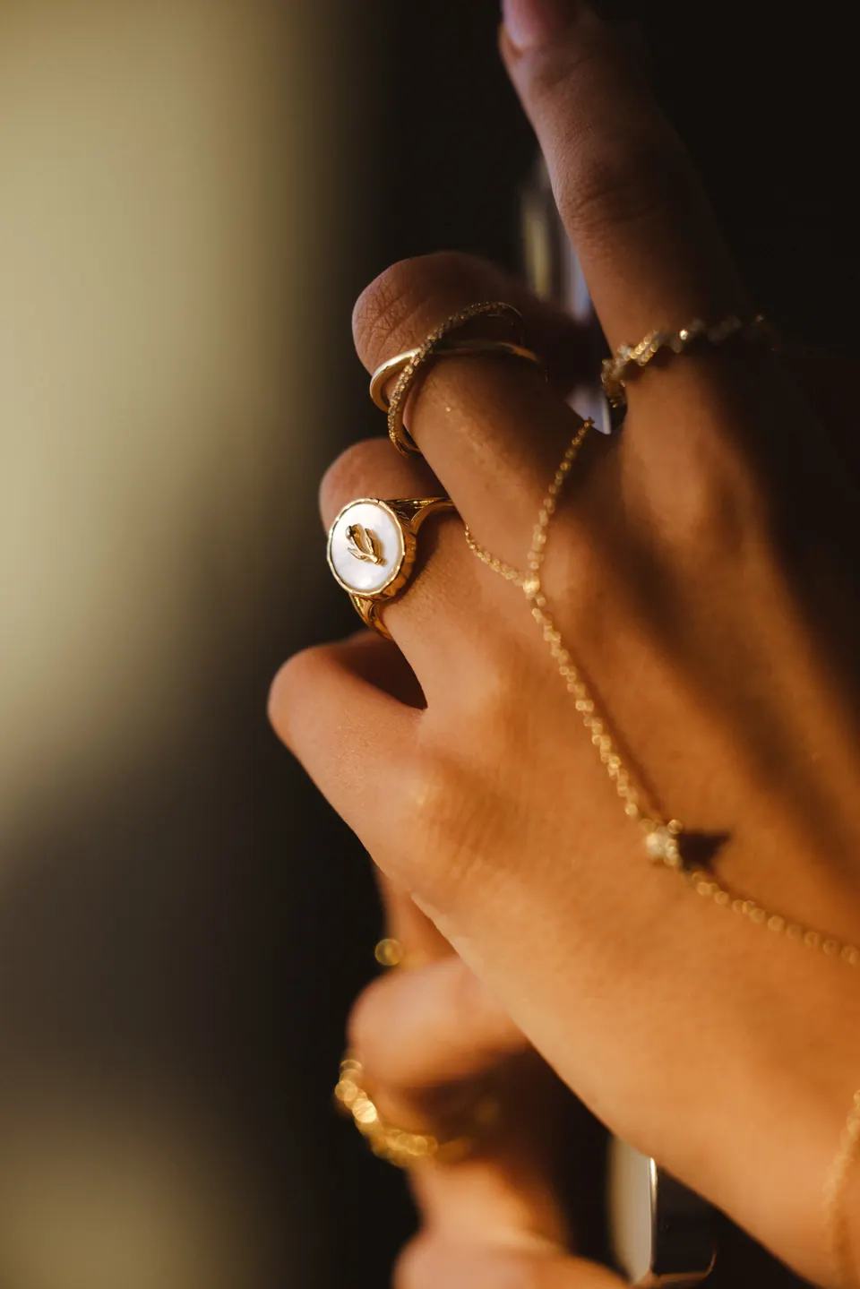 A hand in low warm light wearing a hand chain and a medallion ring, against a dark background.