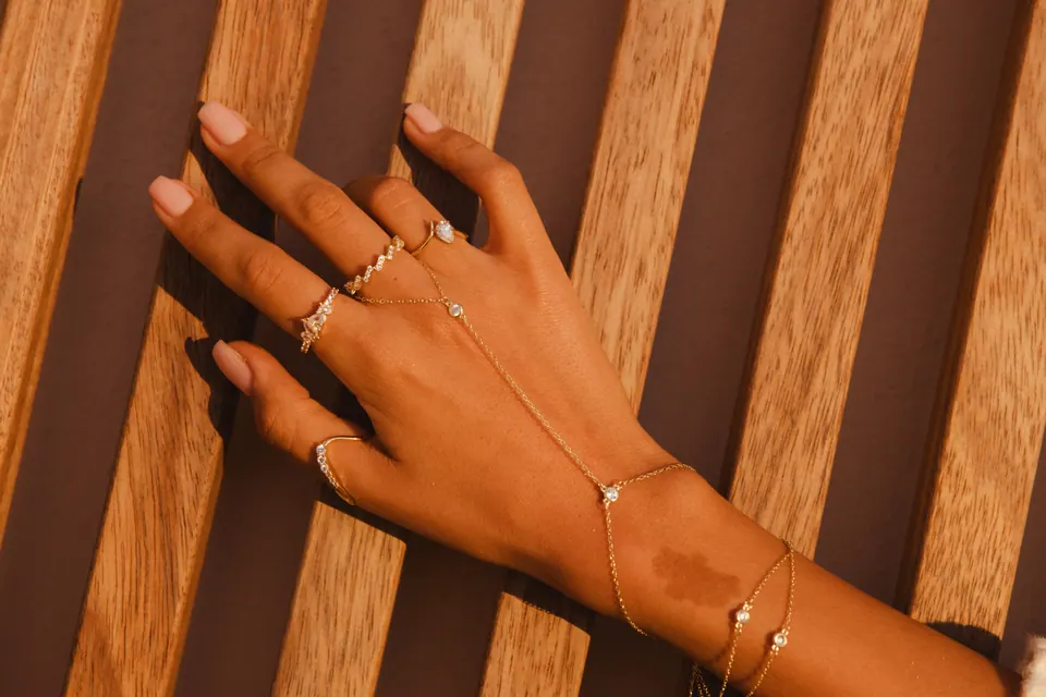 A hand pressed against wooden slats in low golden light, wearing rings and a hand chain, with striped shadows across the skin.