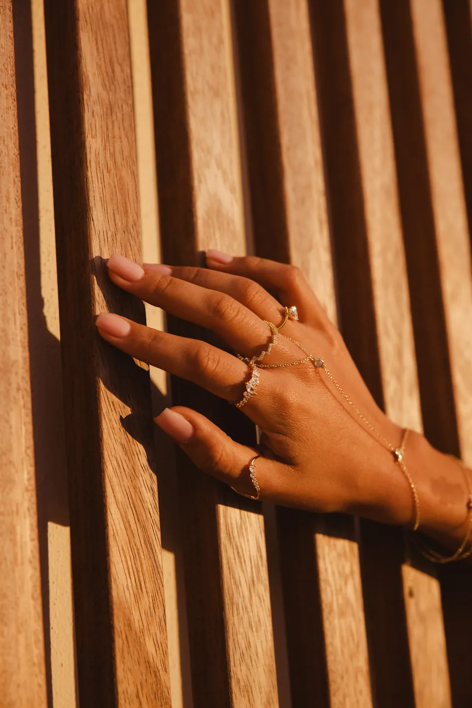 A hand with a fine hand chain resting on wooden slats in low golden light, striped with shadow.
