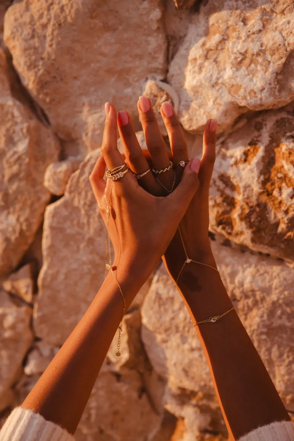 Two raised hands meeting against a sunlit stone wall, wearing rings and a hand chain.