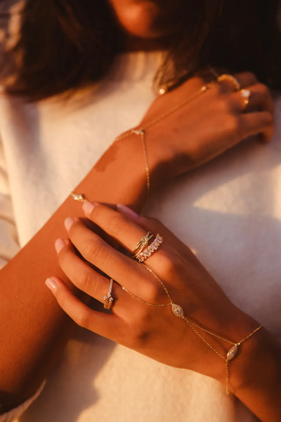 Arms crossed over a white knit jumper in golden light, the hands wearing rings and a hand chain.