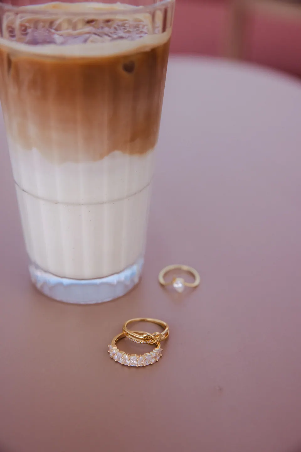 An iced latte on a pink cafe table, with rings resting on the table beside the glass.