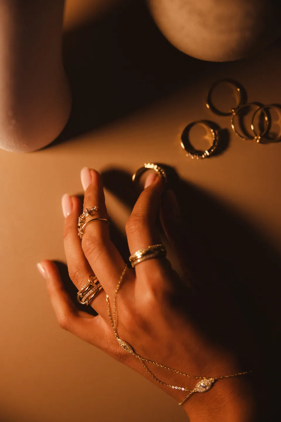 A hand in low, warm light wearing several rings and a hand chain, with more rings on the table behind it.