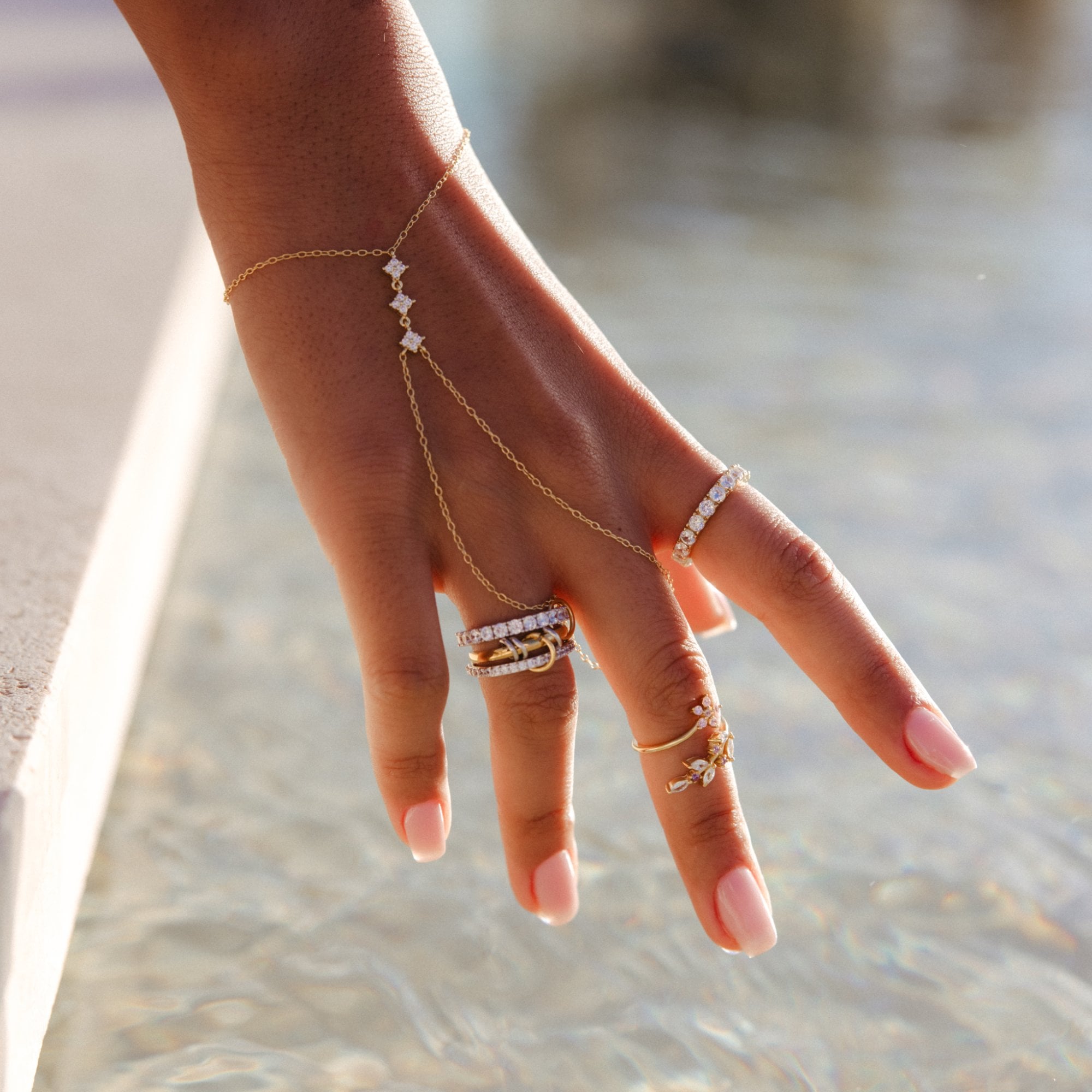 Amal hand chain worn on a hand reaching over clear, sunlit water, with stacked rings.