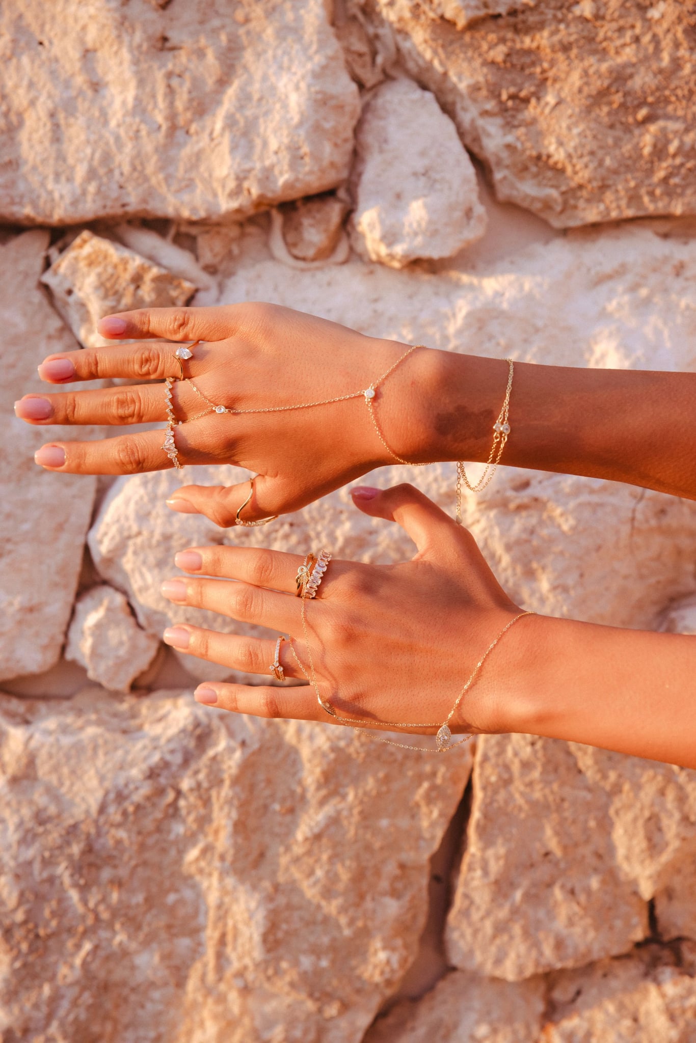Lailah hand chain worn with rings, two hands resting on a pale stone wall in warm light.