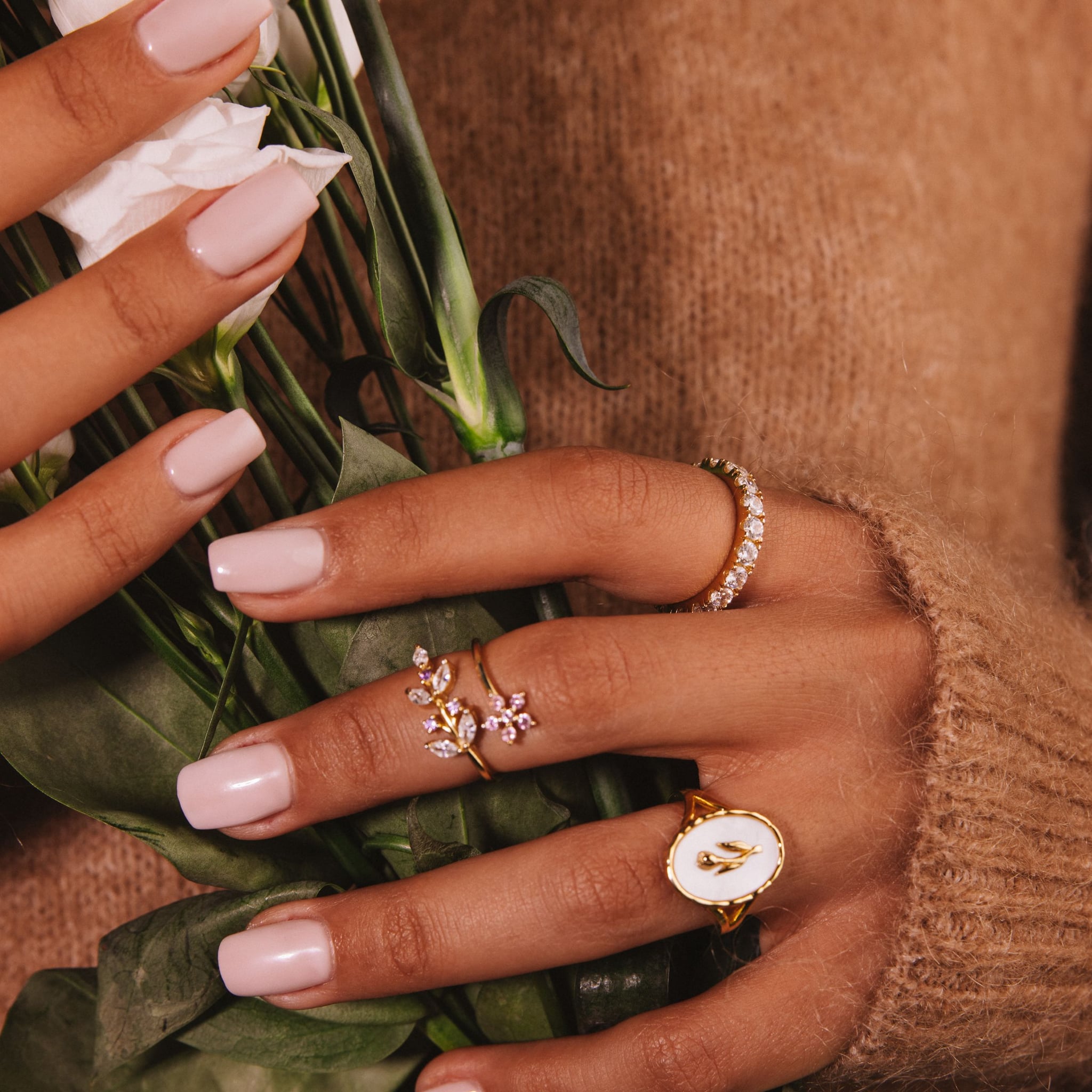 Hands holding white flowers against a camel knit, wearing the Lilly ring and other rings.