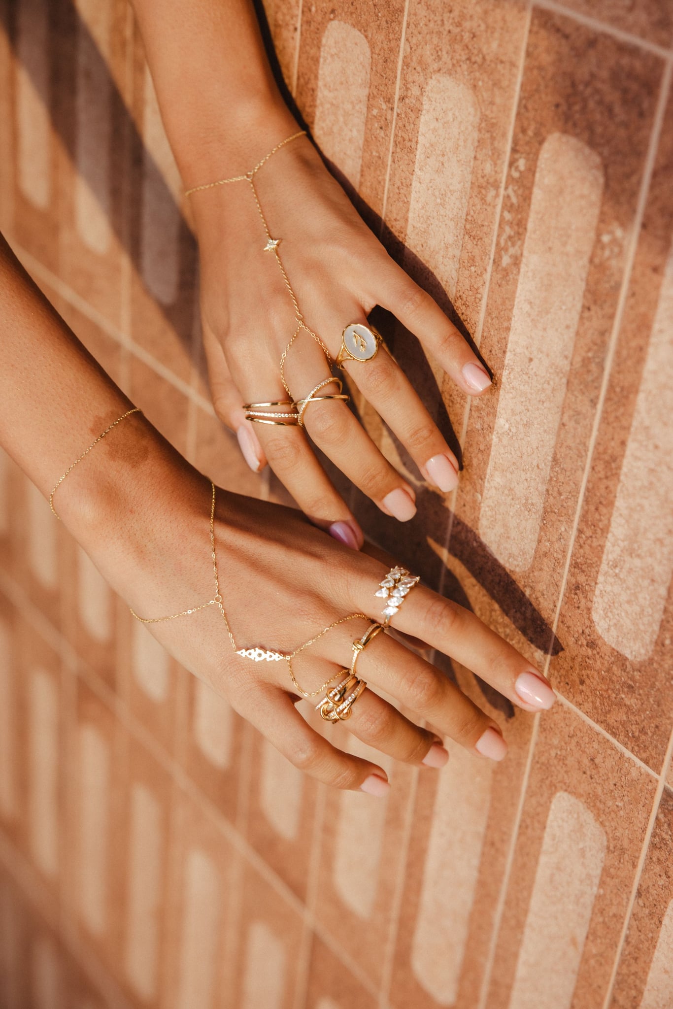 Two hands on terracotta tiles wearing the Nour hand chain and rings.