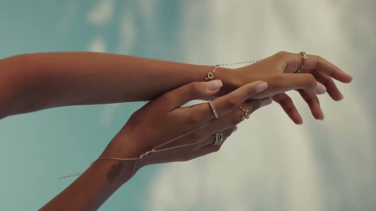 Hands wearing gold-plated rings and a fine hand chain, raised against a pale sky.